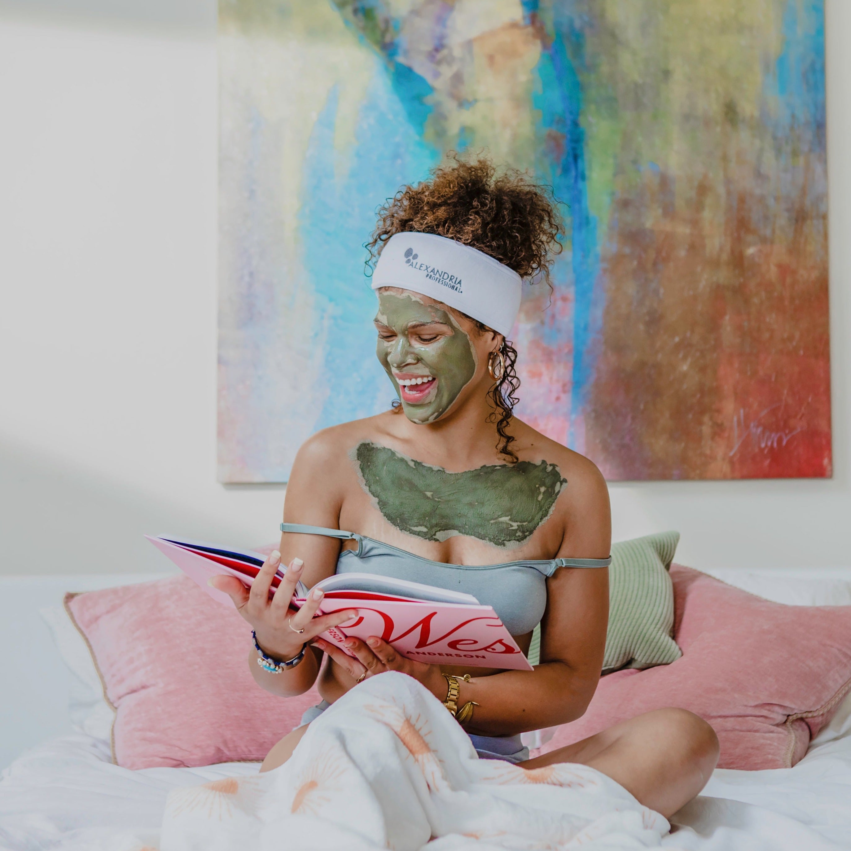 a happy woman sitting on a bed with a green mud mask applied to her face and chest, reading a book.