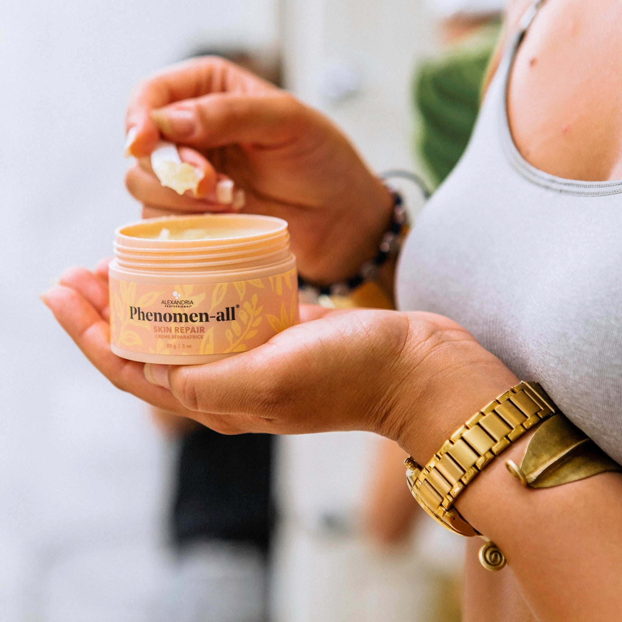 a hand holding a 3 ounce jar of Phenomen-all skin repair cream in their palm, using a mini spoon to remove some product. 